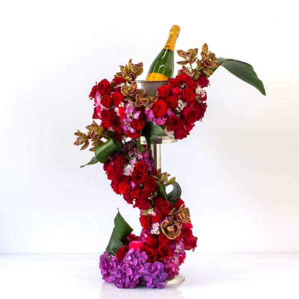 Tall sculptural red and purple floral arrangement spiraling around a stand with a champagne bottle on top