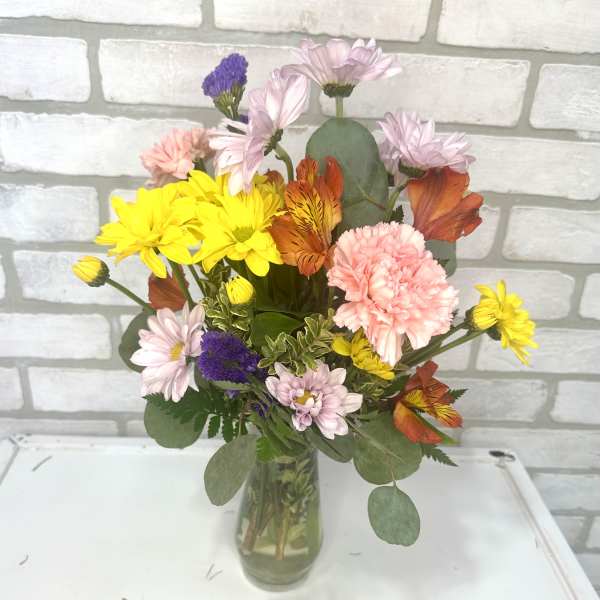 Mixed bouquet of yellow daisies, pink carnations, and orange blooms in a clear glass vase