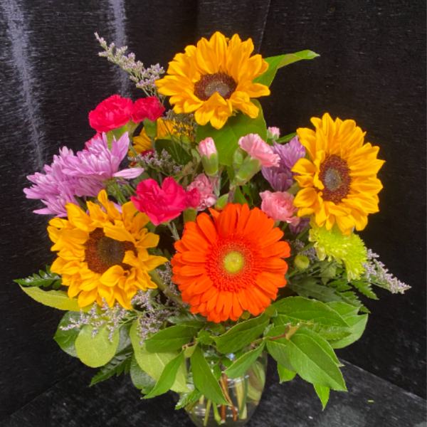 Bright mixed bouquet with sunflowers, orange gerbera, carnations, and mums in a clear glass vase