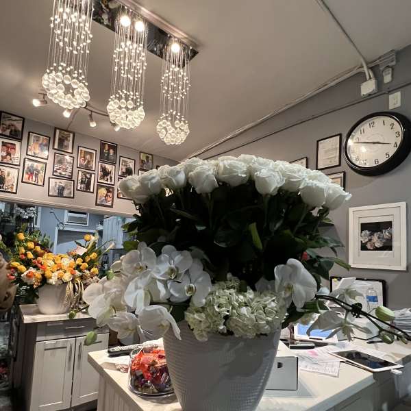 Large white arrangement of roses, orchids, and hydrangea in a white pot on a florist counter