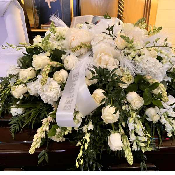 Large white funeral casket spray of roses and mixed blooms with ribbon on a dark wood casket