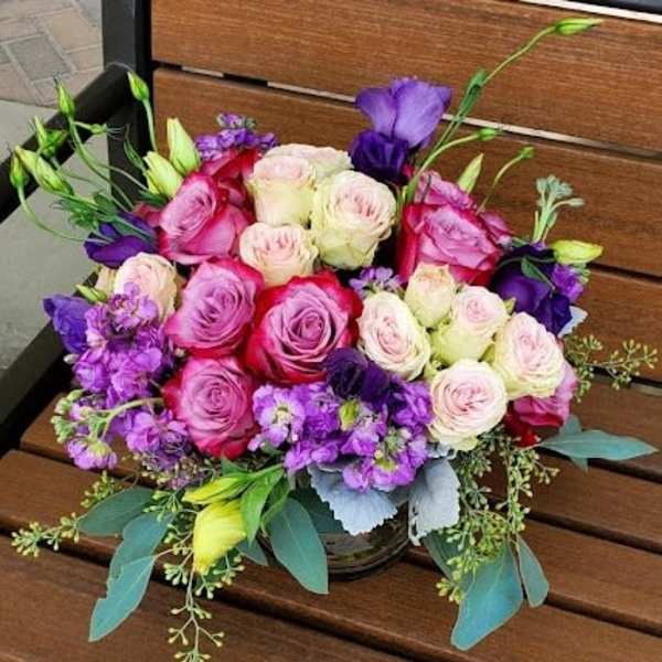 Low arrangement of pink and cream roses with purple flowers in a clear vase on a wooden bench