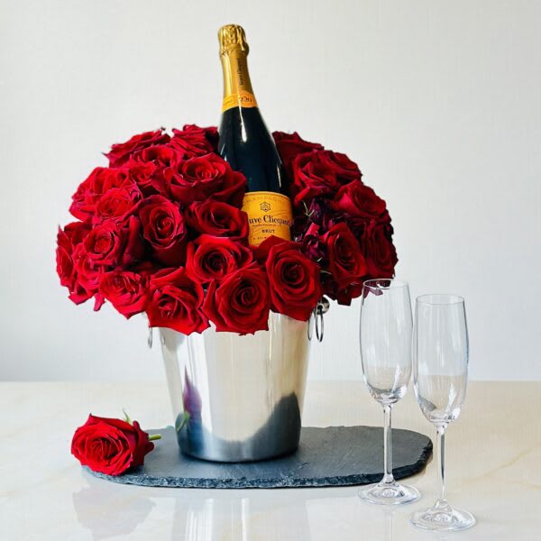 Red roses arranged around a champagne bottle in a silver bucket with two flutes.