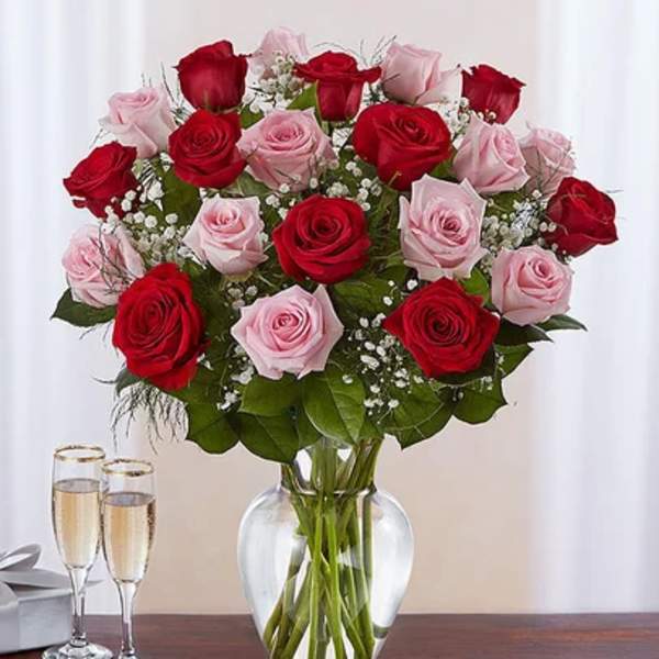 Arrangement of red and pink roses with baby's breath in a clear glass vase beside two champagne flutes.