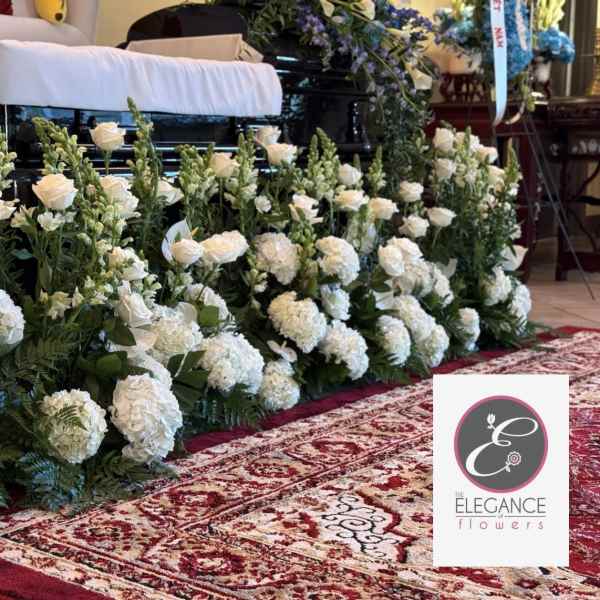 Long row of white roses, hydrangeas, and snapdragons arranged in front of a casket on a red patterned rug
