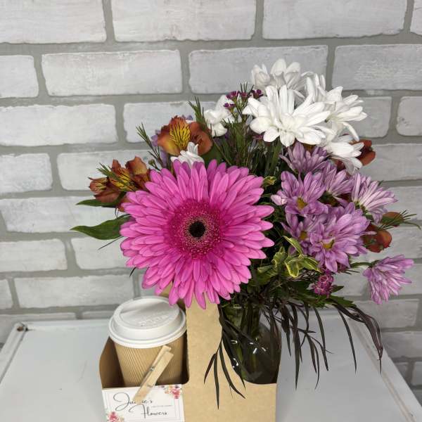 Mixed bouquet with bright pink gerbera daisy in glass vase next to a to-go coffee cup in a cardboard caddy.
