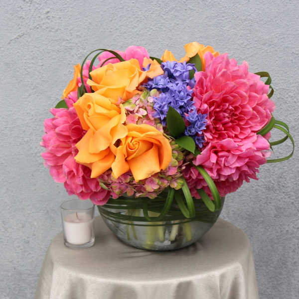 Bright bouquet of orange roses, pink dahlias, and purple flowers in a glass bowl vase