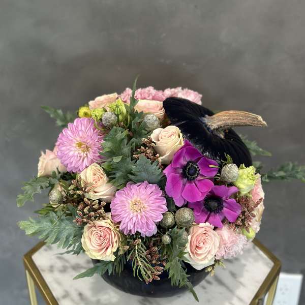 Pink and cream flowers arranged in a black vase with a dark feather accent.