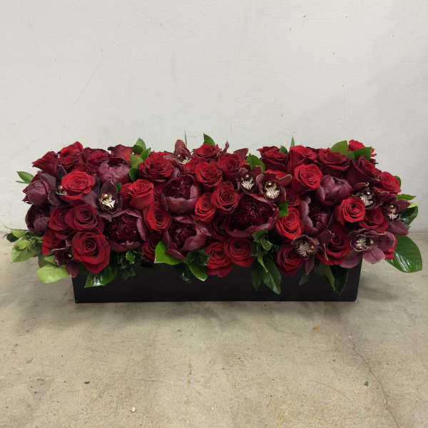 Low rectangular arrangement of dark red roses, peonies, and orchids in a black container