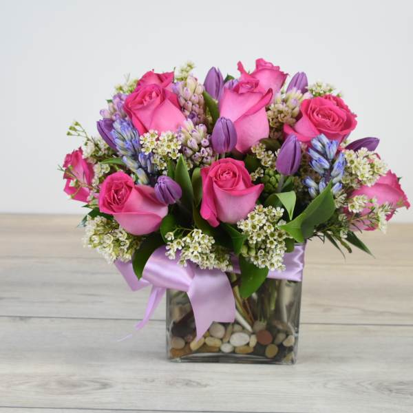 Compact arrangement of hot pink roses with purple blooms in a square glass vase with ribbon.