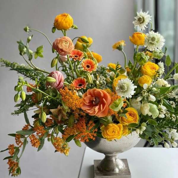 Mixed orange, yellow, white, and peach flowers in a silver pedestal vase