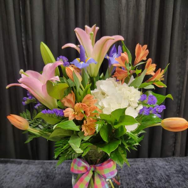 Mixed bouquet of pink lilies, orange blooms, white hydrangea and purple flowers in a glass vase with plaid bow.