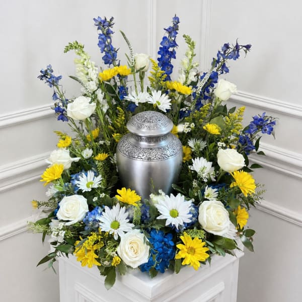 White roses, blue flowers, and yellow daisies arranged around a silver urn