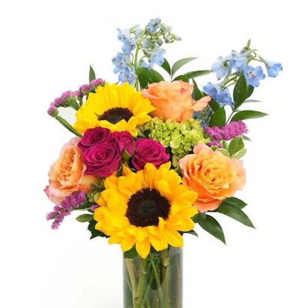 Tall glass vase of yellow sunflowers, peach and magenta roses, blue delphinium and mixed blooms