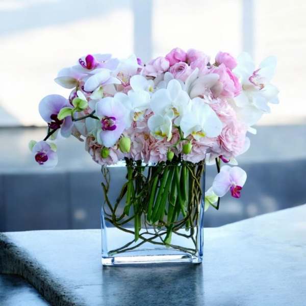 Low square vase of white orchids and soft pink flowers with visible stems.
