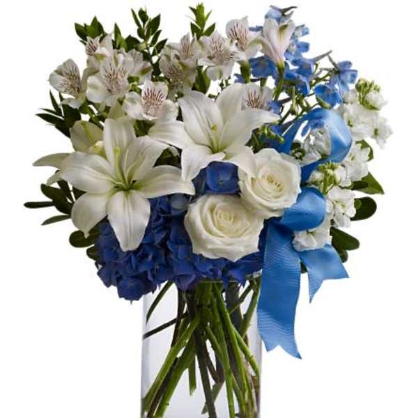 White lilies and roses with blue and white blooms in a clear glass vase with a blue ribbon bow