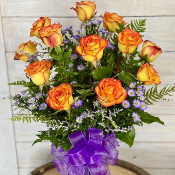 Orange-yellow roses in a glass vase with purple filler flowers and a purple ribbon