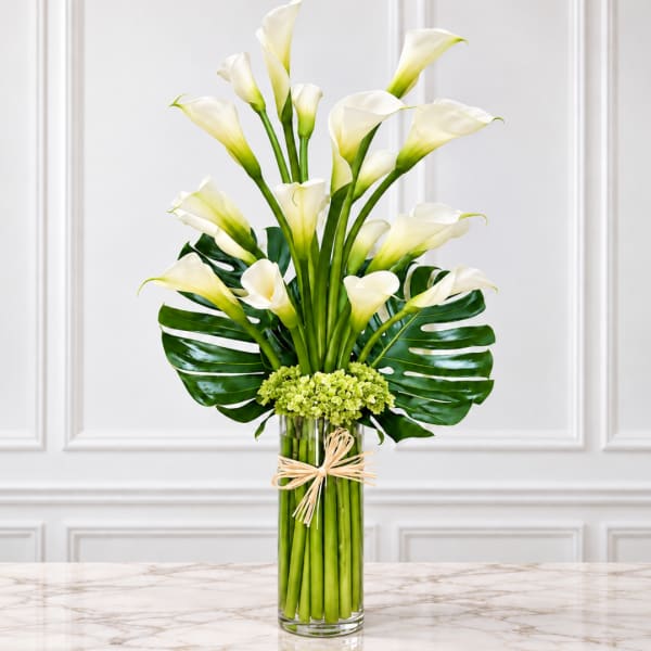 White calla lilies in a tall glass vase with green tropical leaves