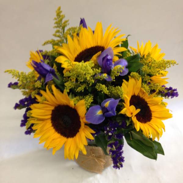 Arrangement of bright yellow sunflowers with purple irises in a glass vase wrapped in burlap.