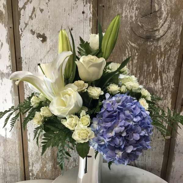 White lilies and roses with a blue hydrangea cluster in a silver vase.
