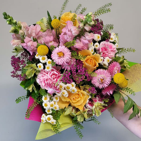 Hand-tied bouquet of pink and yellow flowers including roses, snapdragons, carnations, and daisies in bright wrap.