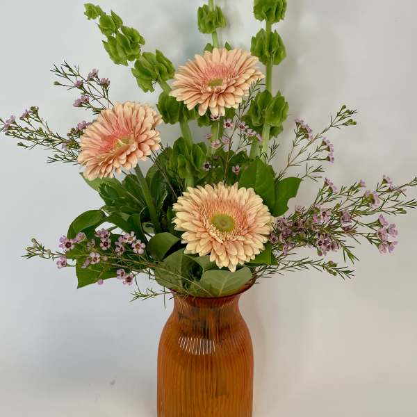 Peach gerbera daisies in an amber vase with green spikes and small purple flowers