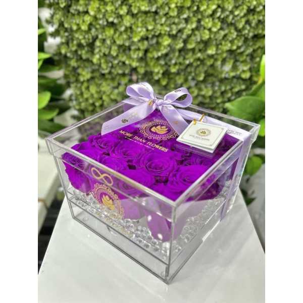 Square acrylic box filled with vivid purple roses, topped with a lavender bow and small tag.