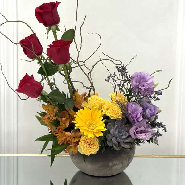 Mixed floral arrangement with red roses, yellow blooms, and purple flowers in a gray bowl vase