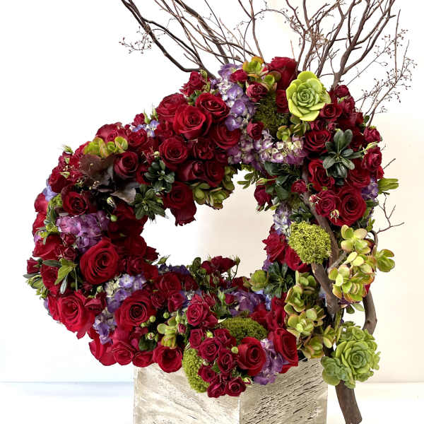Circular arrangement of red roses, purple blooms, and green succulents with branches in a rustic square container