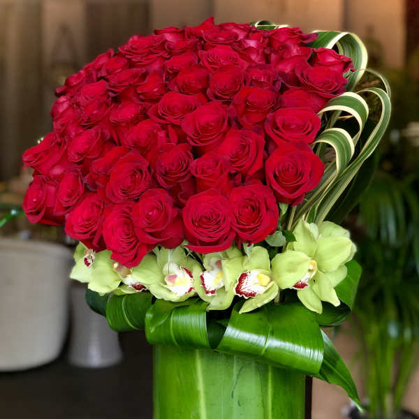Tall arrangement of red roses with green cymbidium orchids in a leaf-lined green glass vase