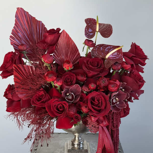 Deep red rose arrangement in a silver urn with burgundy tropical accents