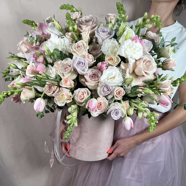 Large pastel arrangement of roses, tulips, and orchids in a blush hatbox held by a person