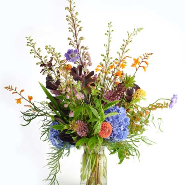 Tall mixed arrangement with blue hydrangeas, orange blooms, and wildflowers in a clear glass vase