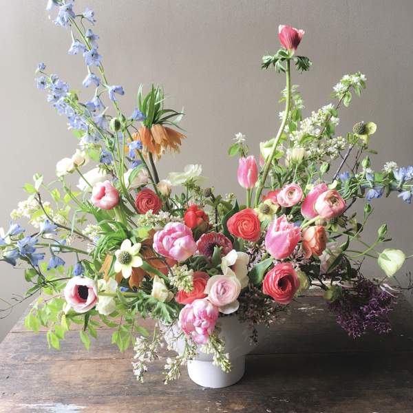 Asymmetrical garden-style arrangement of pink, coral, and white flowers with blue accents in a white vase