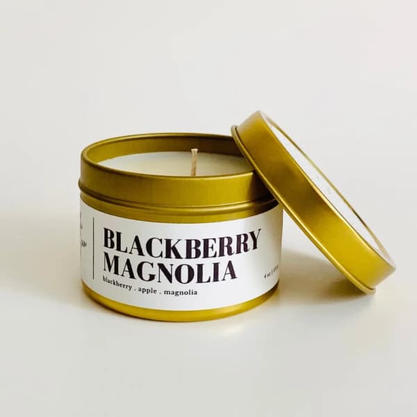 Blackberry Magnolia candle in a gold tin with lid leaning beside it