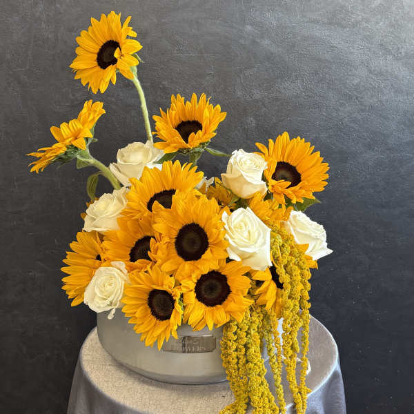 Sunflowers and white roses arranged in a gray hatbox with trailing yellow blooms.