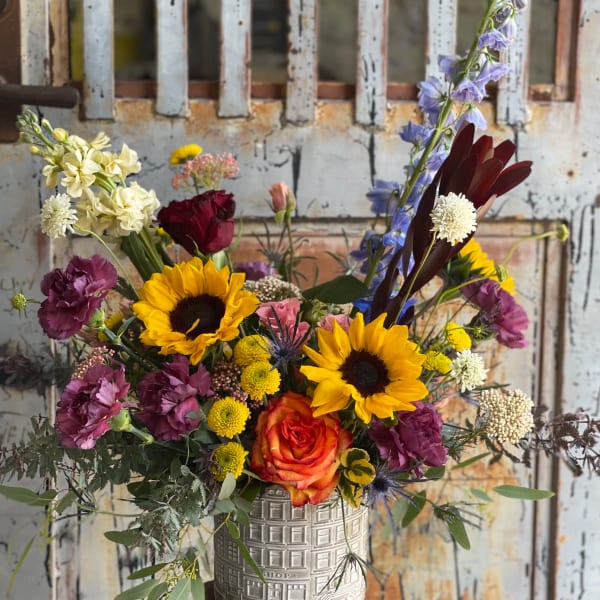 Mixed bouquet with sunflowers, roses, and purple blooms in a decorative metal container