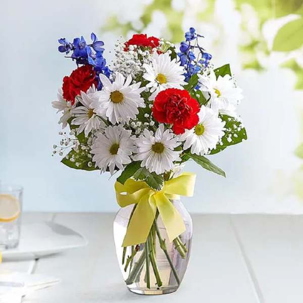 Bouquet of white daisies, red carnations, and blue flowers in a glass vase with a yellow ribbon