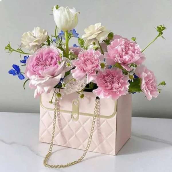 Pink and white flowers arranged in a quilted handbag-shaped box with a chain strap.