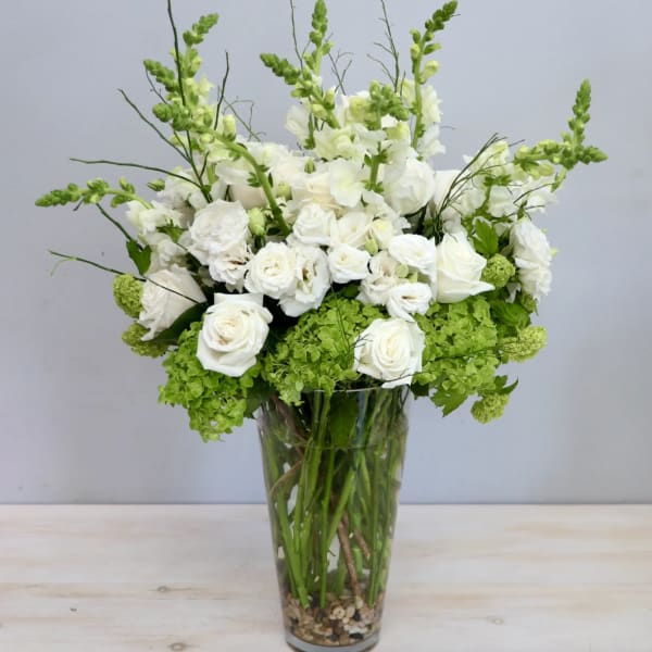 Tall arrangement of white roses and snapdragons with green hydrangeas in a clear glass vase with pebbles