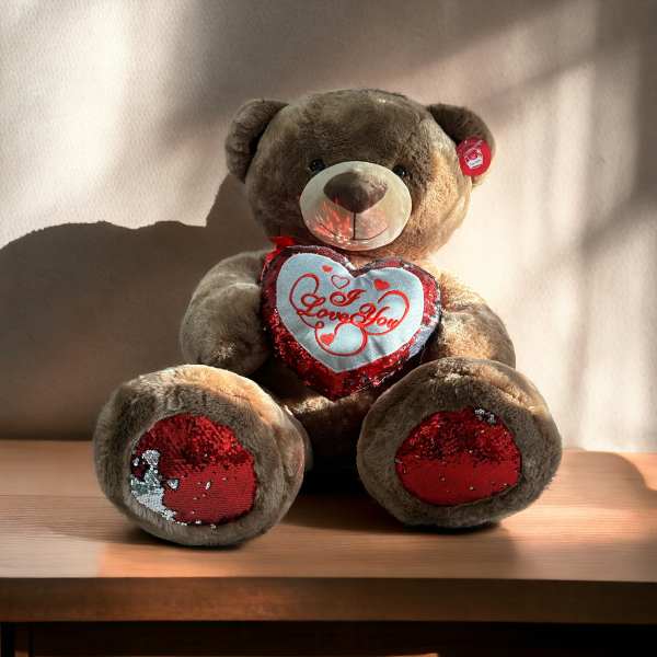 Brown teddy bear with red sequin heart reading "I Love You" on a wooden surface