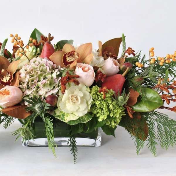 Low rectangular centerpiece with peach roses, orchids, hydrangea, berries, and evergreen accents in a glass vase