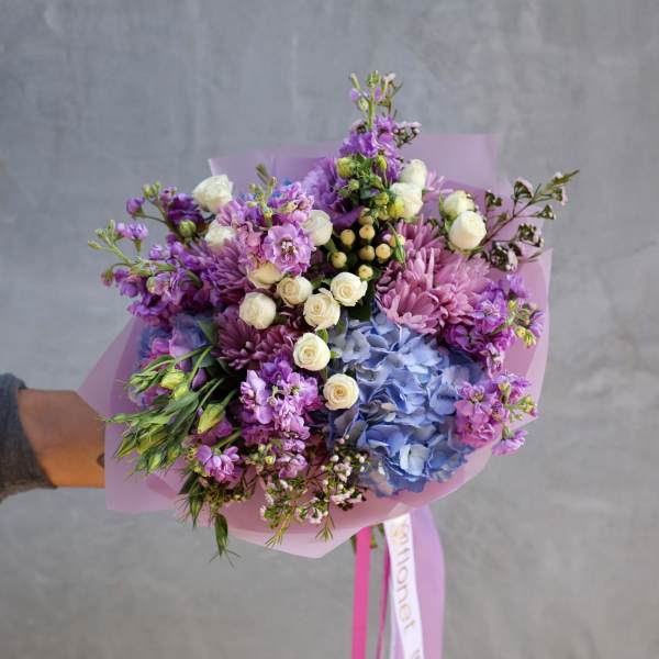 Handheld bouquet of purple, blue, and white flowers wrapped in pink paper