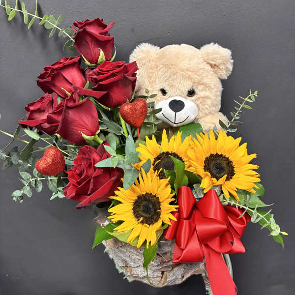Red roses and sunflowers with a teddy bear and red ribbon in a rustic container