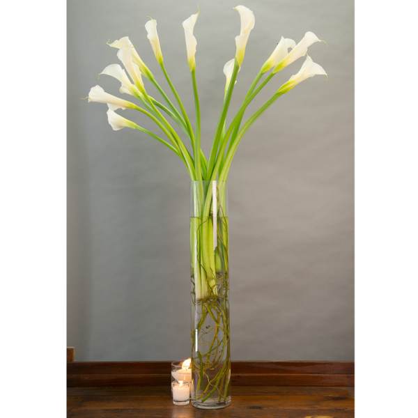 White calla lilies in a tall glass vase with candles beside it