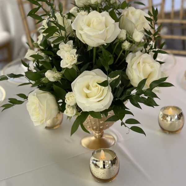 White roses arranged in a gold vase on a table with small candles