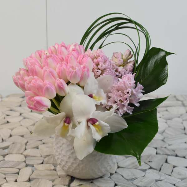 Compact arrangement of pink tulips, white orchids, and pastel hyacinths in a white textured vase with looping green blades.