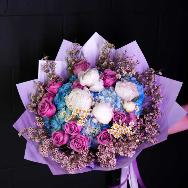 Bouquet of pink roses, white peonies, and blue hydrangeas wrapped in lavender paper