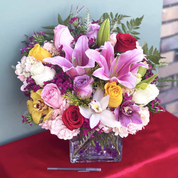 Mixed bouquet of pink lilies, roses, and orchids in a square glass vase