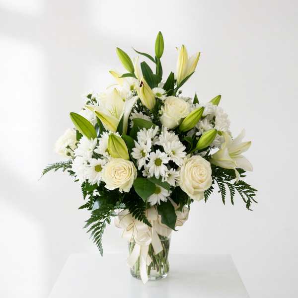 White lilies, roses, and daisies arranged in a glass vase with a ribbon
